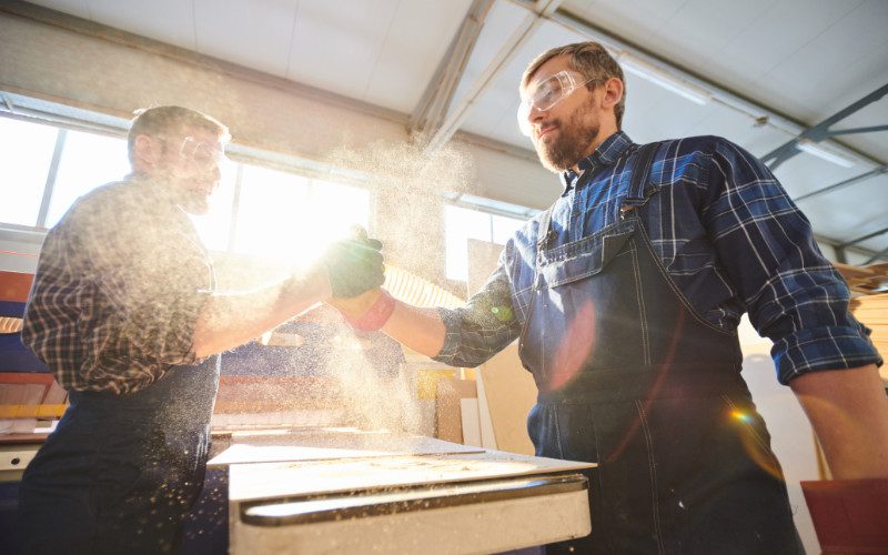 Two manufacturers shaking hands with a cloud of dust surrounding their hands. They both have goggles on and are wearing checkered shirts.
