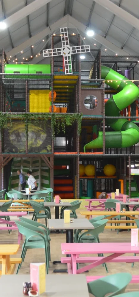 This is the second part of the main Brocksbushes image. It features the main view of the softplay, including features like the green tube slide, white windmill and green/white chevron slide. You can also see pink, green and yellow tables and chairs in front of the softplay.