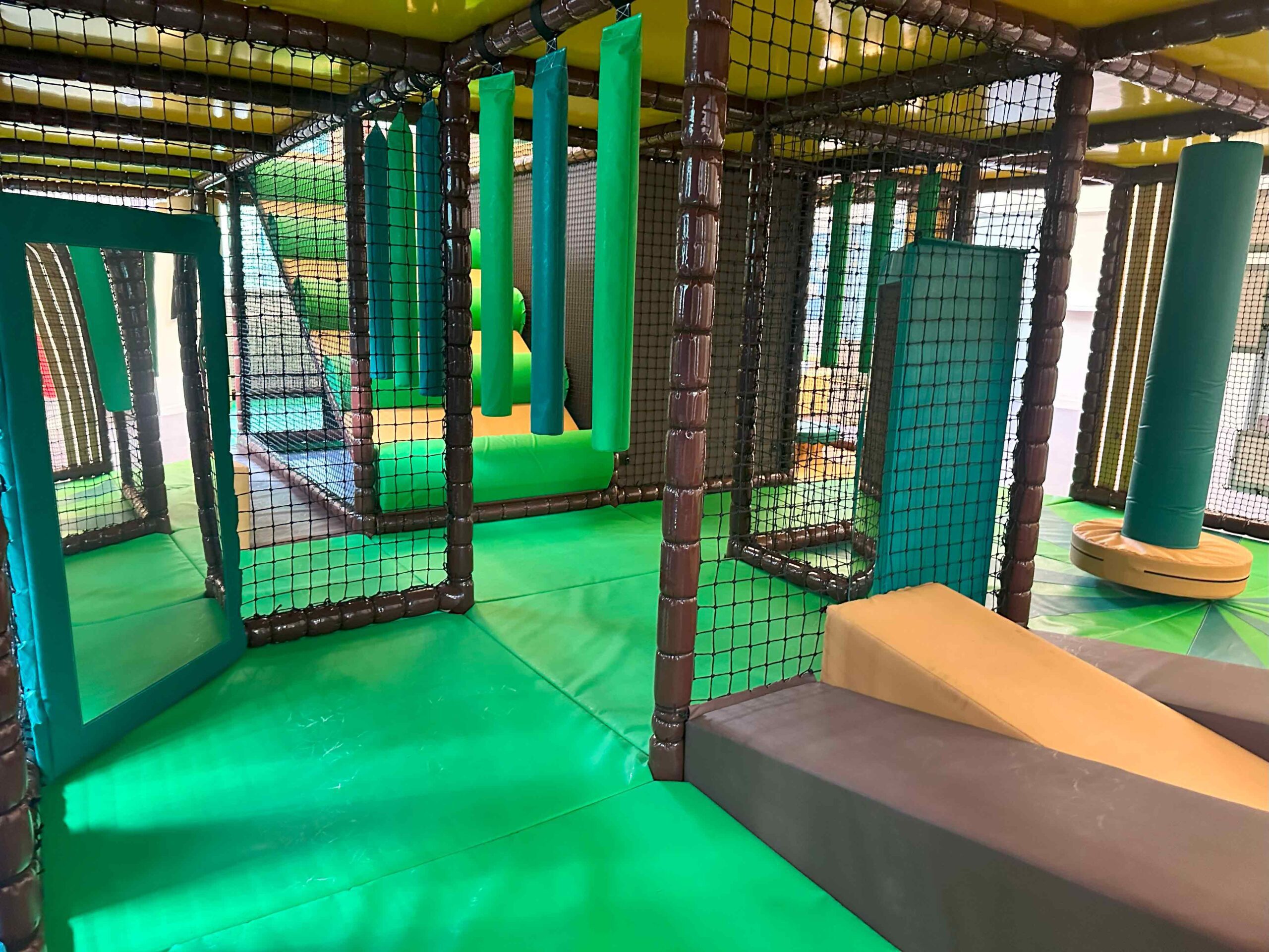 Inside view of the brown, green and orange theme softplay. A green and orange dizzy disc, orange and brown wedge floor and mirrors. There are also some green hanging snakes. All can be found at the Awesome CIC's indoor softplay area designed by Play Creations.