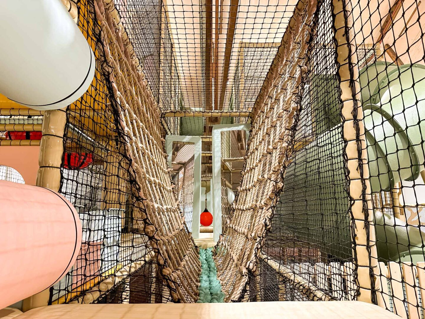 The Play Hut's indoor softplay area's personalised V-net bridge. It's a beige and blue net bridget next to the green spiral tube slide. This was designed by Play Creations