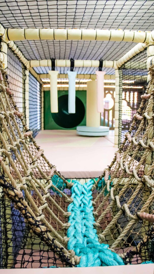 Blue and cream rope bridge leading across the softplay area. In the background there are 3 hanging snakes and a dizzy disc. Found at Cub and Cup.